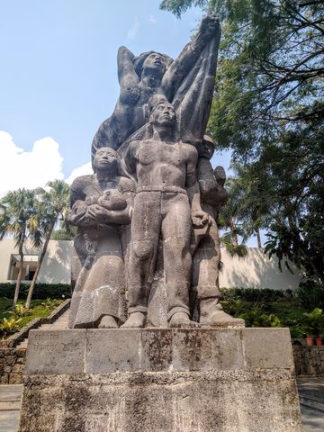 A large stone sculpture of several human figures stands on a pedestal in a park. Museum of Art of El Salvador, Antiguo Cuscatlan, El Salvador, January 30, 2020
