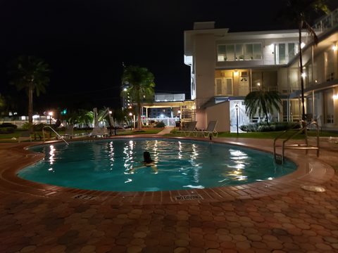 A person swims in a lit outdoor pool at night next to a multi-story building. Holiday Isle Yacht Club, Sunrise, USA, October 11, 2020