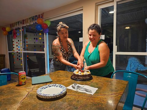 Two women are smiling while cutting a chocolate birthday cake together at a table. MJ, Casa Del Mar Esmeralda, Chelem, Mexico, April 5, 2023
