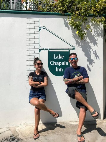A man and a woman pose smiling in front of a white wall with a large green sign. MJ and Cisco, March 6, 2019