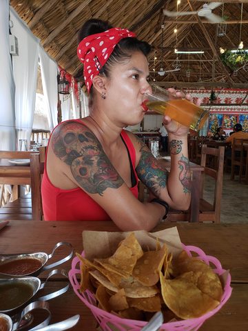 A woman with tattoos and a red polka dot headband drinks from a glass of beer at a wooden table. MJ, Tizimin, Mexico, December 8, 2019