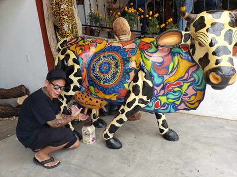 A person in black clothing poses next to a large, brightly painted cow sculpture. Cisco, Artesanías Lucía Sosa, San Francisco Javier, Mexico, October 27, 2019