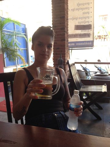 A woman sits at an outdoor table holding a glass of beer and a water bottle. MJ, Central Hostel, Siem Reap, Cambodia, January 24, 2016